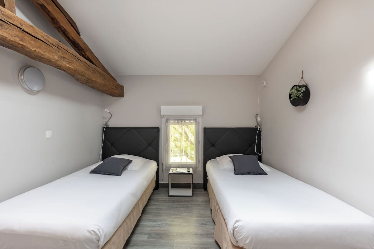 Un dormitorio con dos camas, una ventana, una mesita y una planta colgante.