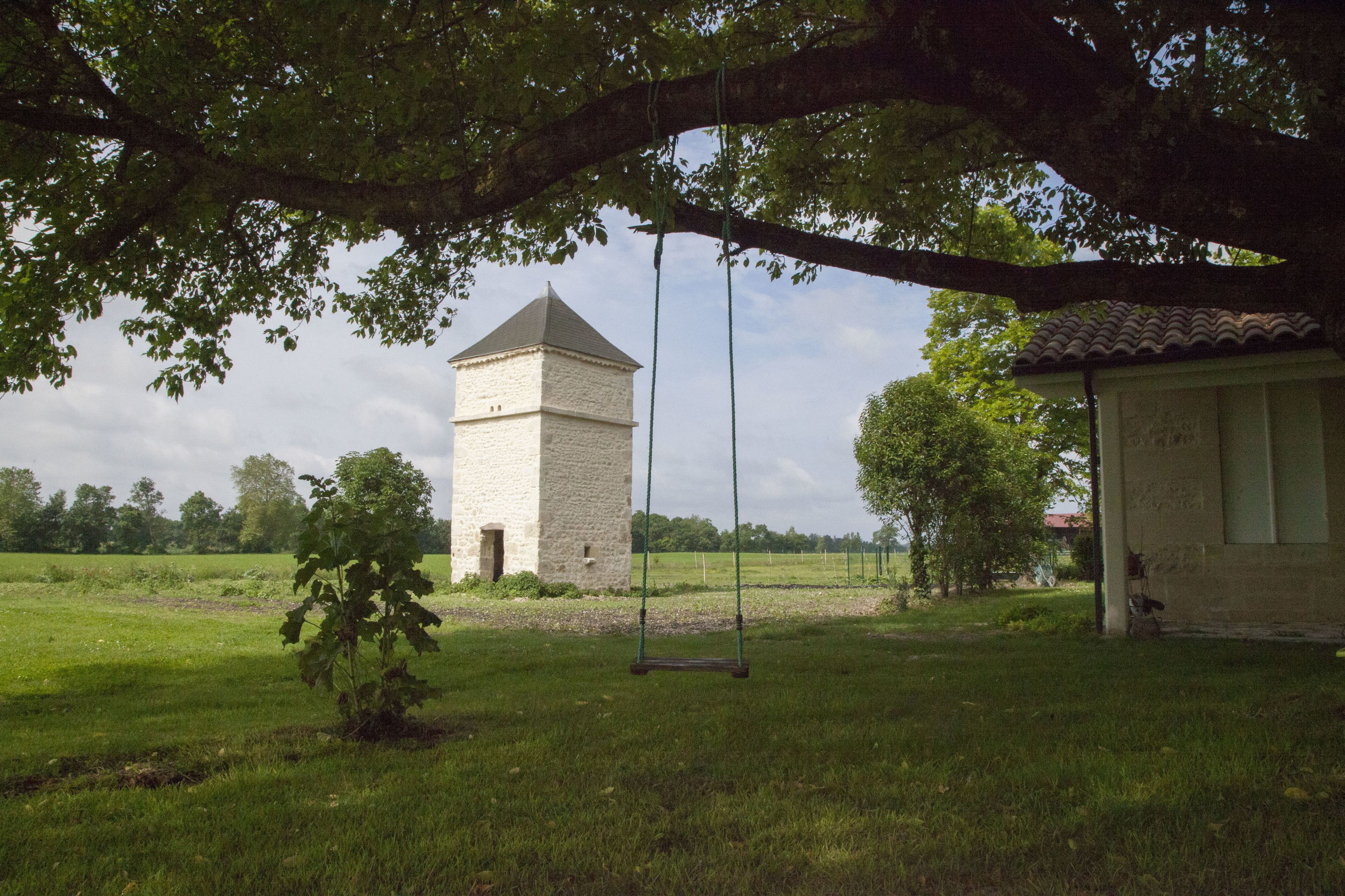 A rustic stone building with a tower, a swing set, and a small tree in a grassy field.