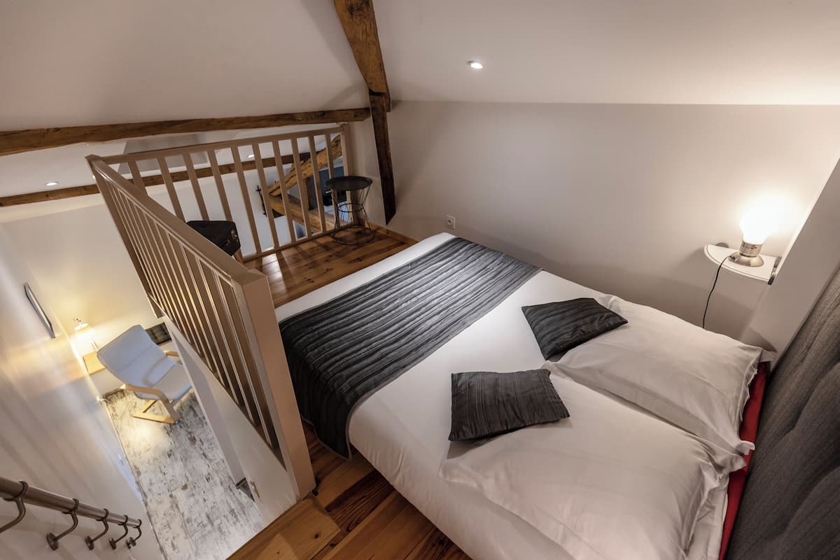 Un dormitorio con una escalera de madera, una cama con ropa de cama blanca y gris, una mesita de noche con una lámpara y una pequeña mesa con una silla.