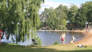 A lakeside park with a swimming area, a grassy area with a chair, and trees. - Evedals Camping (Växjö)