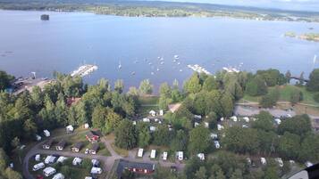 Vue aérienne d’un terrain de camping au bord d’un lac, avec des camping-cars et des tentes, entouré d’une forêt dense et d’un port de plaisance avec des bateaux.