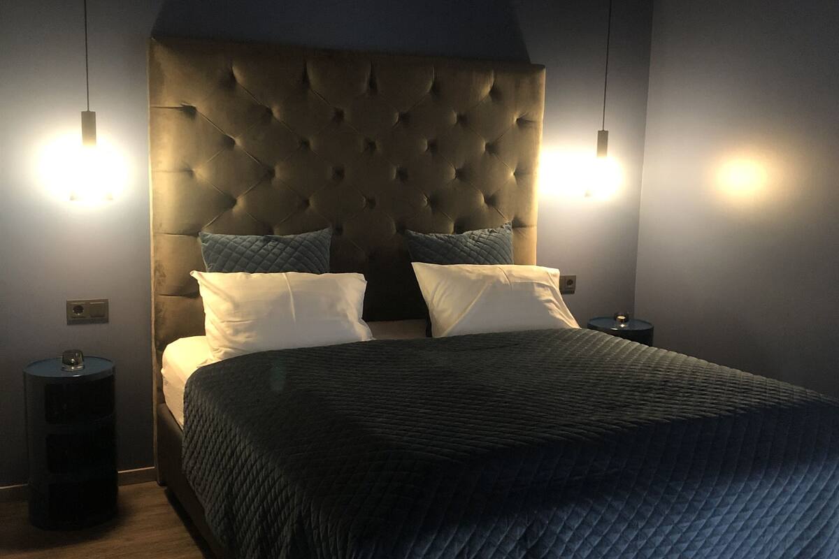 Une chambre à coucher avec une tête de lit capitonnée, deux lampes de chevet, une couverture de lit matelassée de couleur sombre et une petite table de chevet.