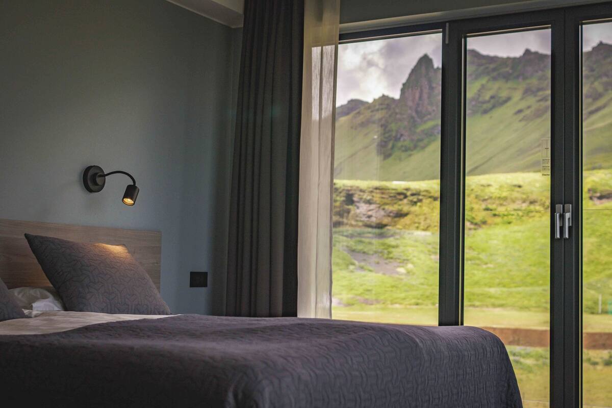 Une chambre avec un grand lit, une table de chevet, une lampe fixée au mur et une vue sur un paysage montagneux, visible à travers une porte-fenêtre coulissante.