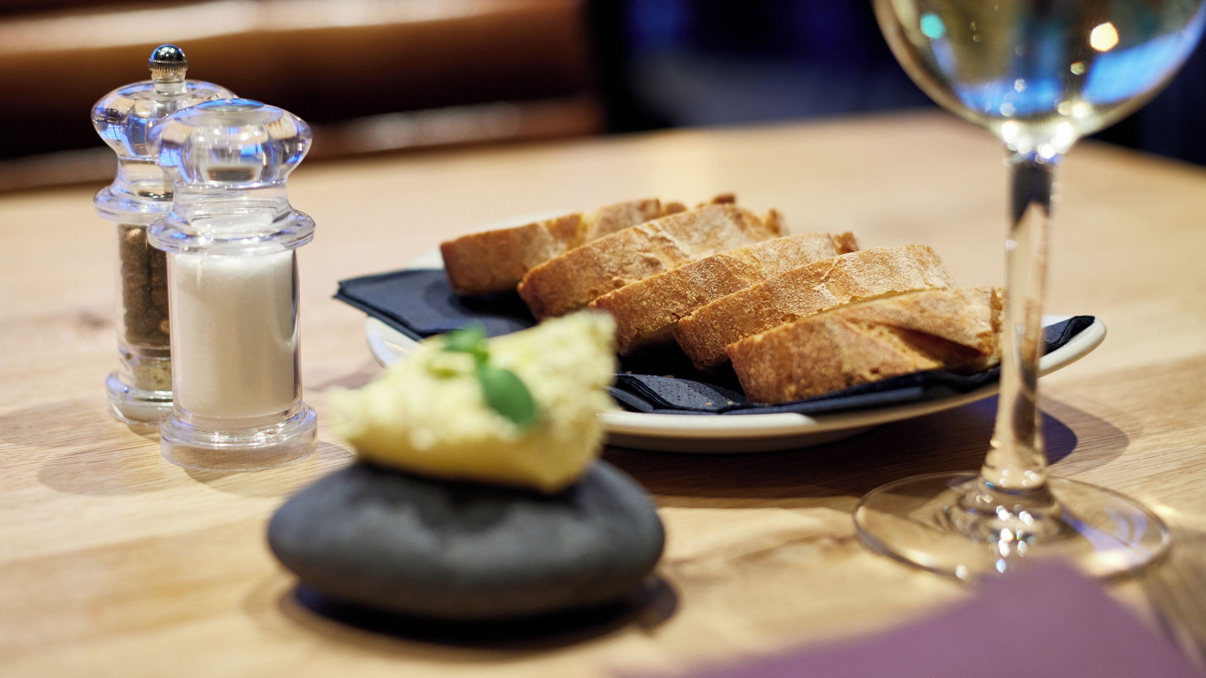 A table with a plate of bread, a salt and pepper shaker, a small bowl of butter, and a wine glass.