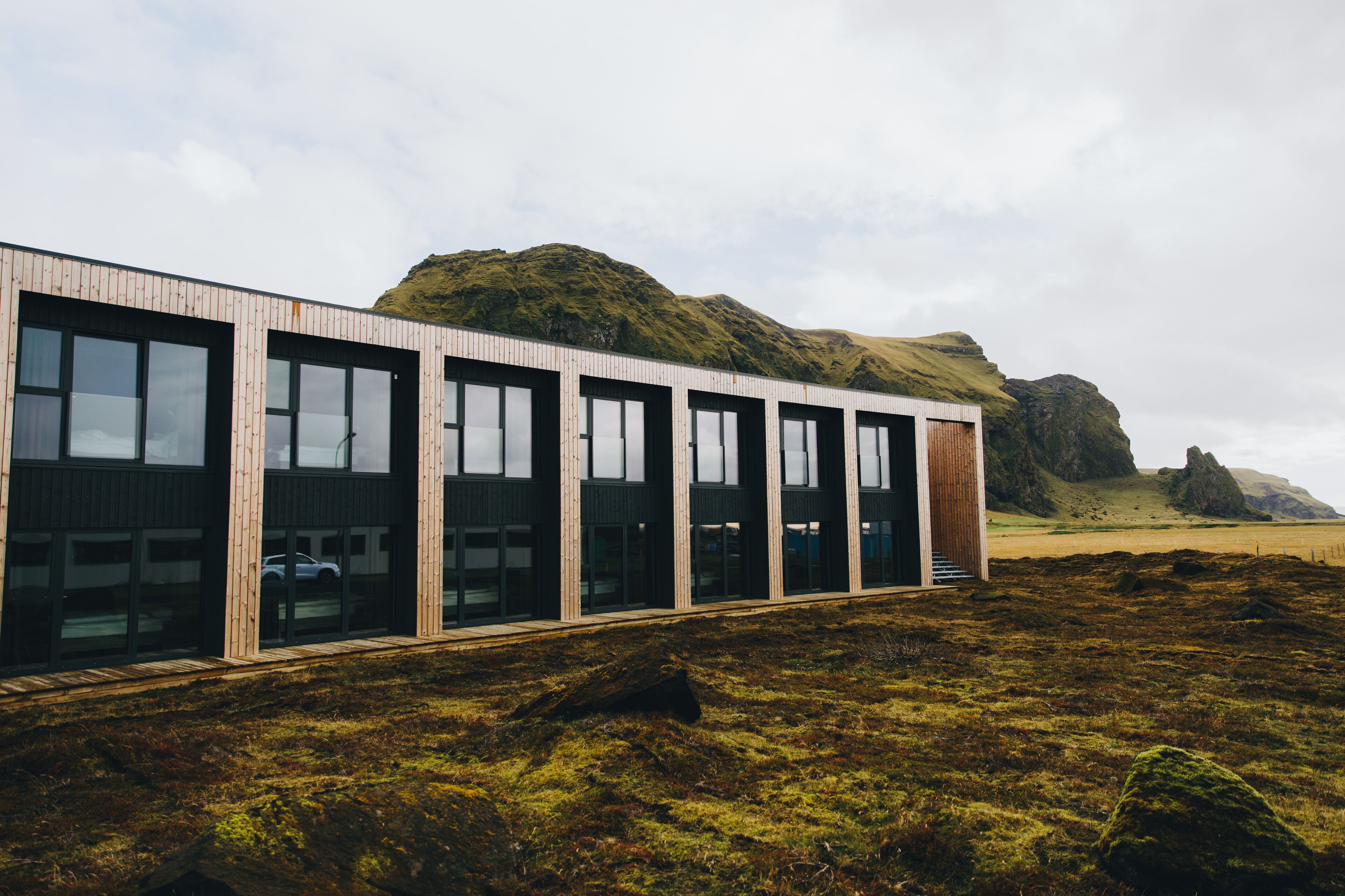A modern building with large windows and a wooden facade, set against a backdrop of green hills and a cloudy sky.