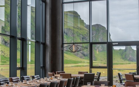 A restaurant with large windows offering a view of green hills. Tables are set with glasses and cutlery.