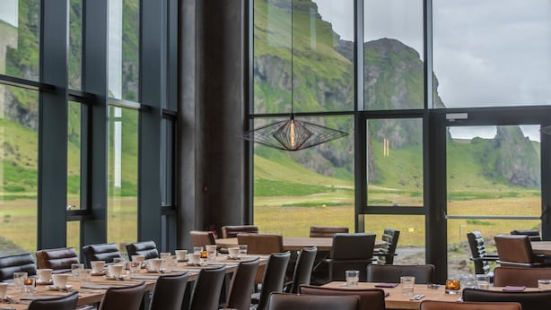 A restaurant with large windows offering a view of green hills. Tables are set with glasses and cutlery. - Hótel Kría (Vik I Myrdal)