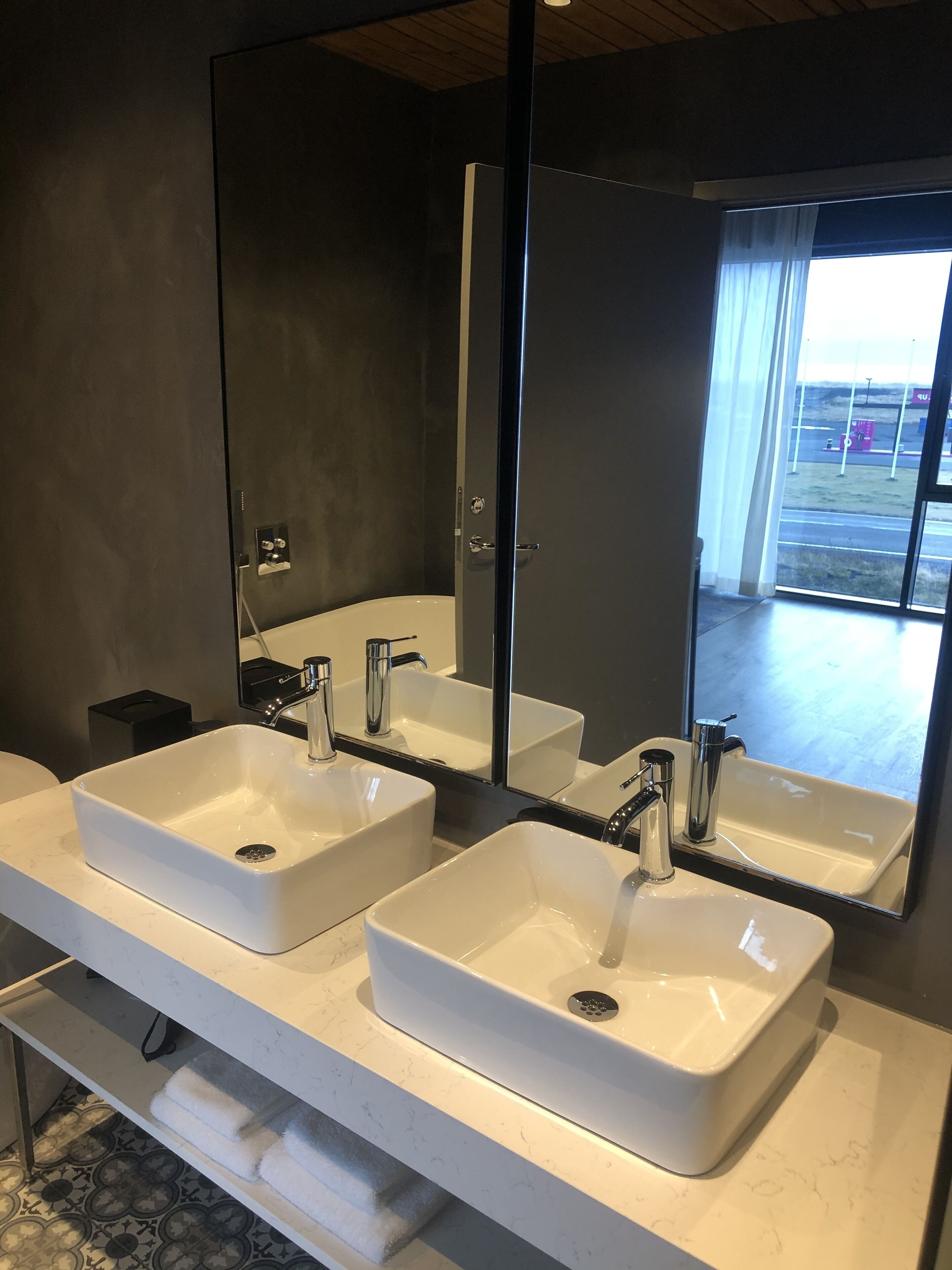 Un baño moderno con dos lavamanos blancos, un espejo grande y vistas al exterior a través de un ventanal.