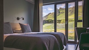 A bedroom with two beds, a large window with a mountain view, and a chair. - Hótel Kría (Vik I Myrdal)