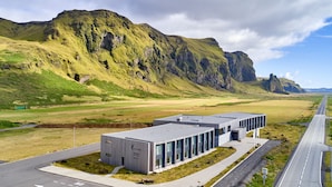A modern hotel with a flat roof and large windows, situated in a scenic area with a road and mountains in the background. - Hótel Kría (Vik I Myrdal)