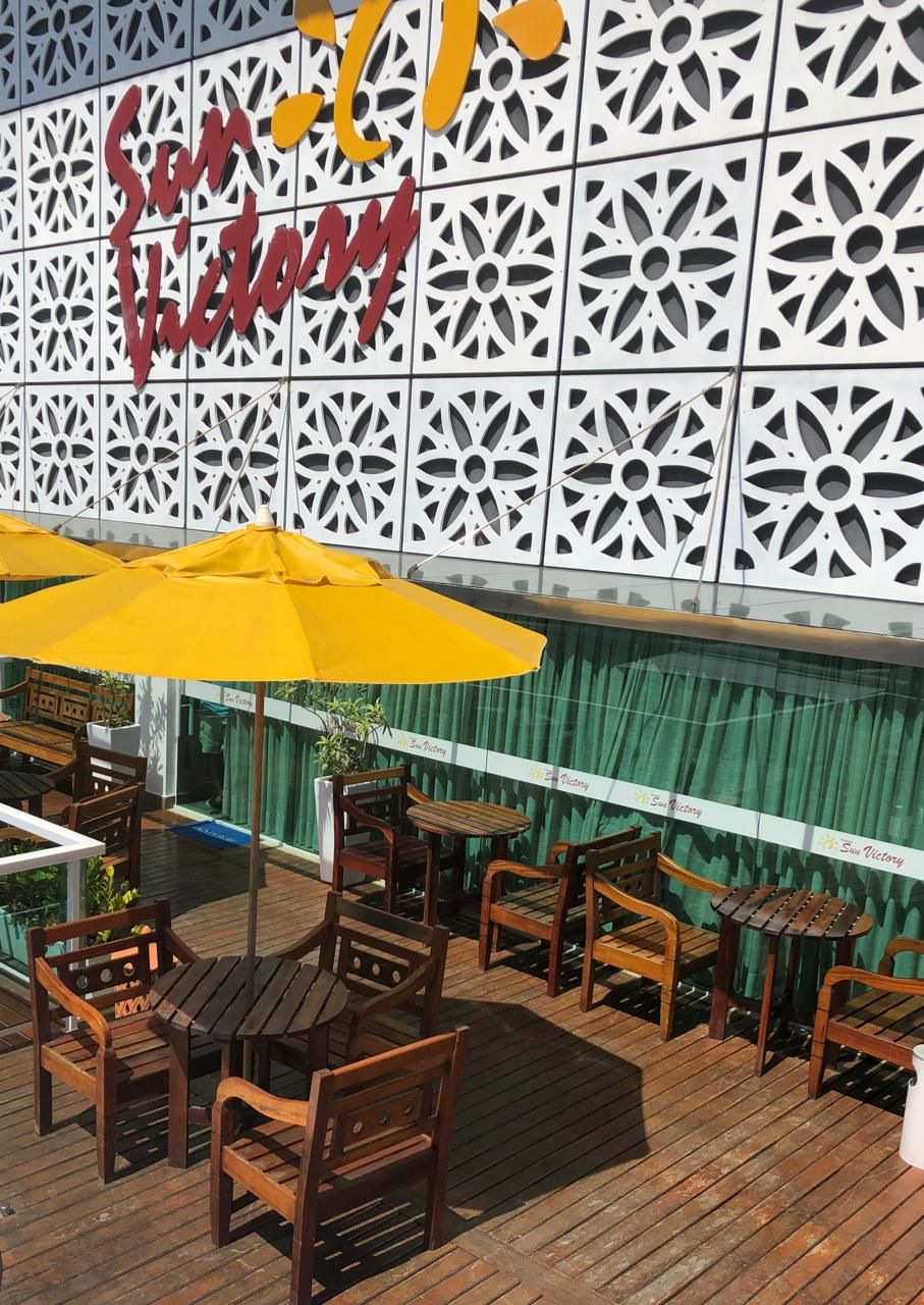 An outdoor seating area with wooden tables and chairs, a yellow umbrella, and a decorative wall with the text 'Sun Victory.'