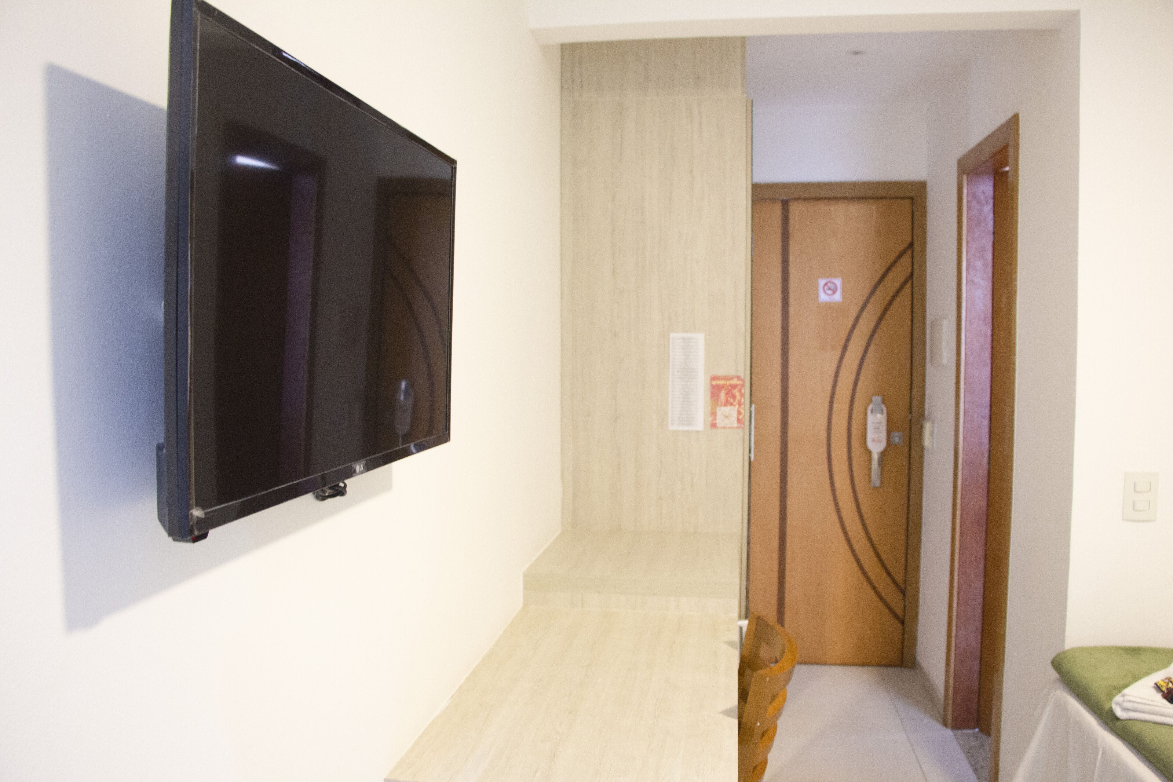 Un couloir avec un téléviseur à écran plat fixé au mur, une porte en bois avec une pancarte et une petite table recouverte d’une nappe verte.