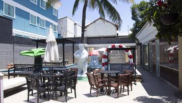 Une terrasse extérieure avec des tables et des chaises, une statue blanche et un parasol rouge et blanc à rayures.