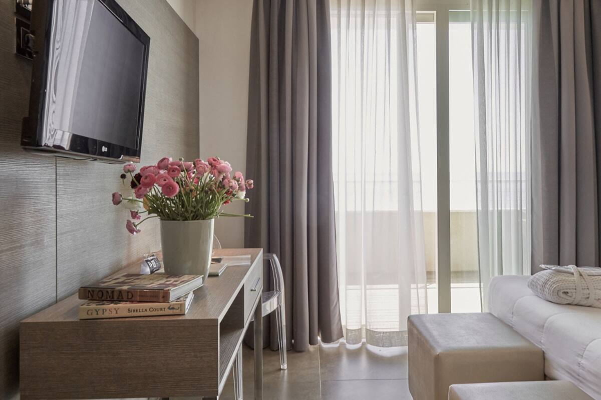 Ein Hotelzimmer mit Bett, Schreibtisch mit Blumenvase, Flachbildfernseher und einem Fenster mit Vorhängen.