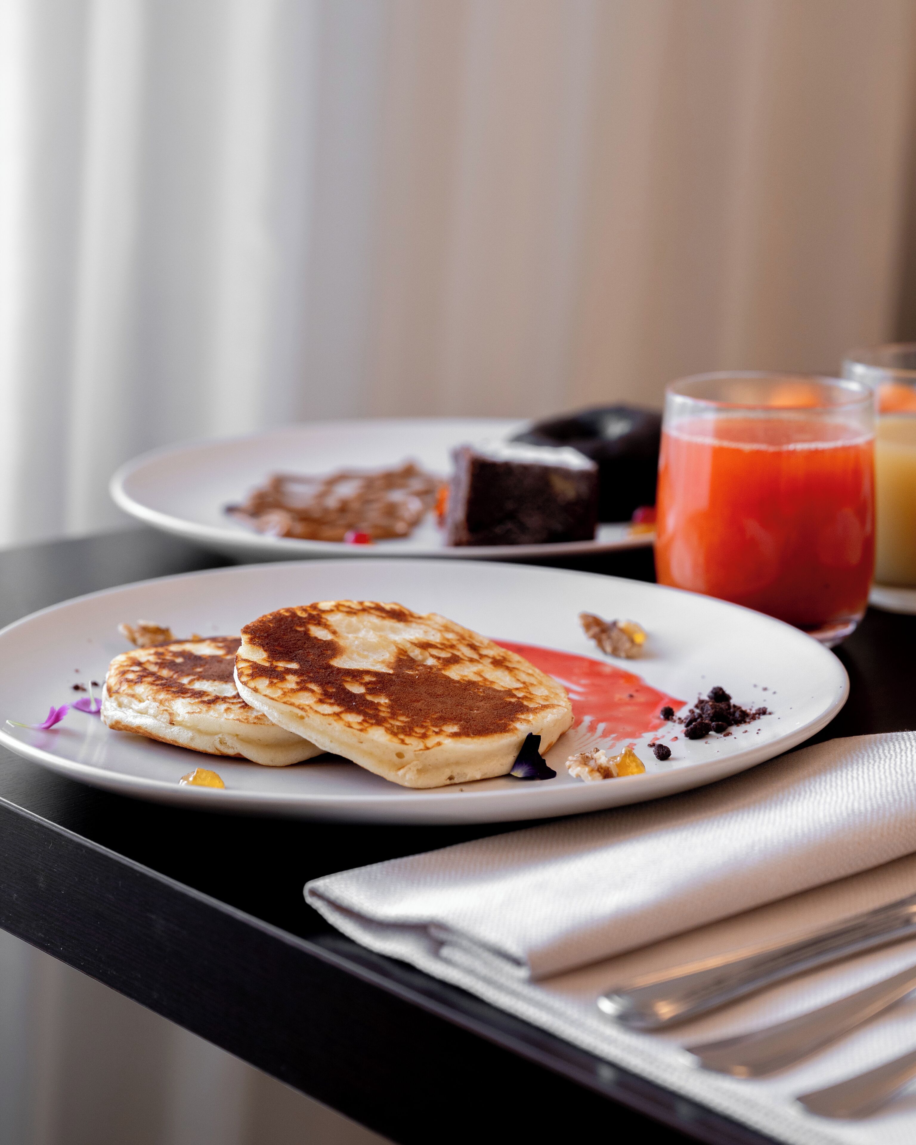 Een ontbijttafel met pannenkoeken op een bord, een glas rode sappen een bord met dessert.