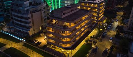 A multi-story hotel with a rooftop terrace, a swimming pool, and a well-lit parking area.