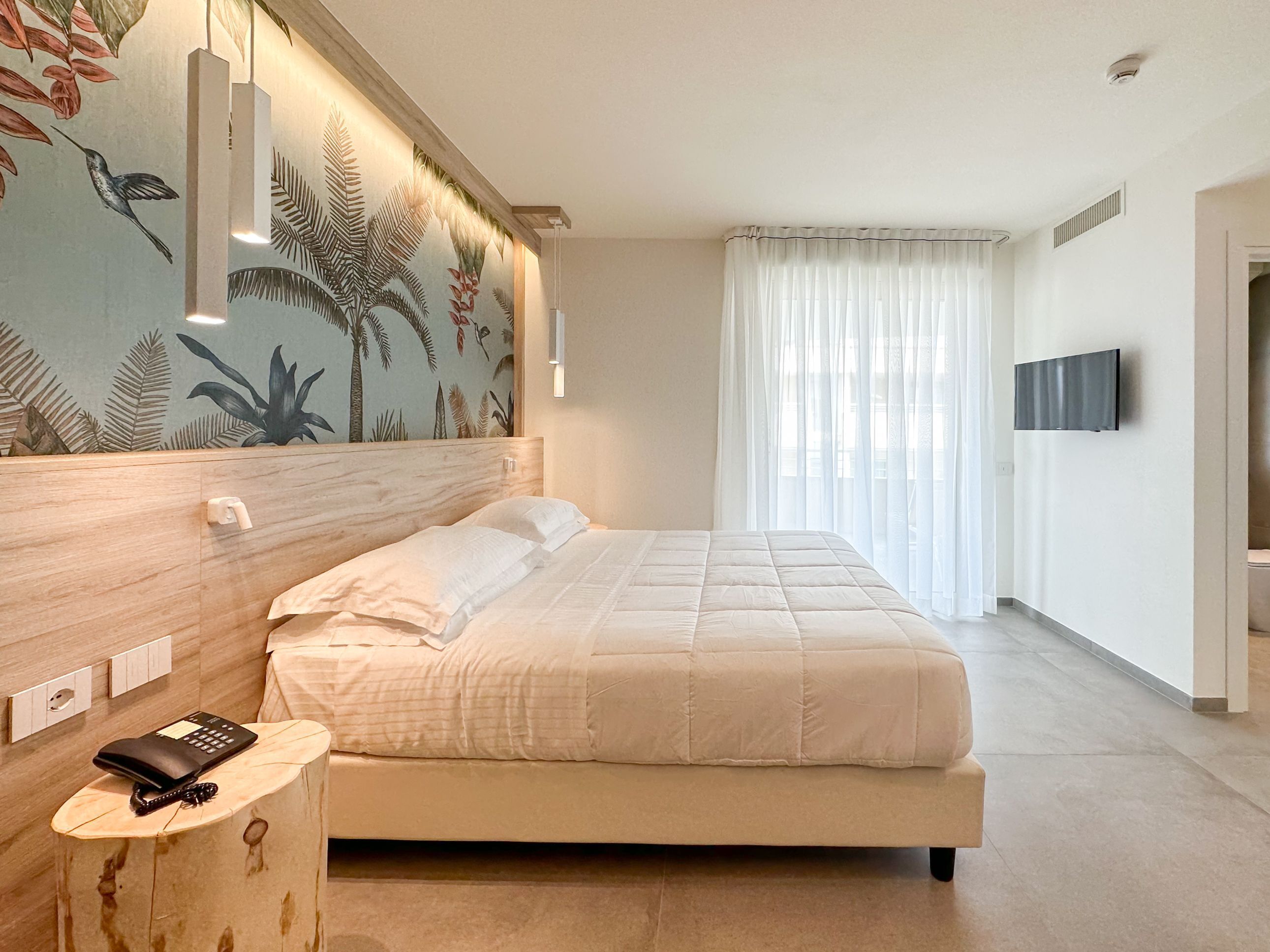 A modern hotel room with a large bed, a wooden nightstand, a mounted TV, and a tropical-themed wall mural.
