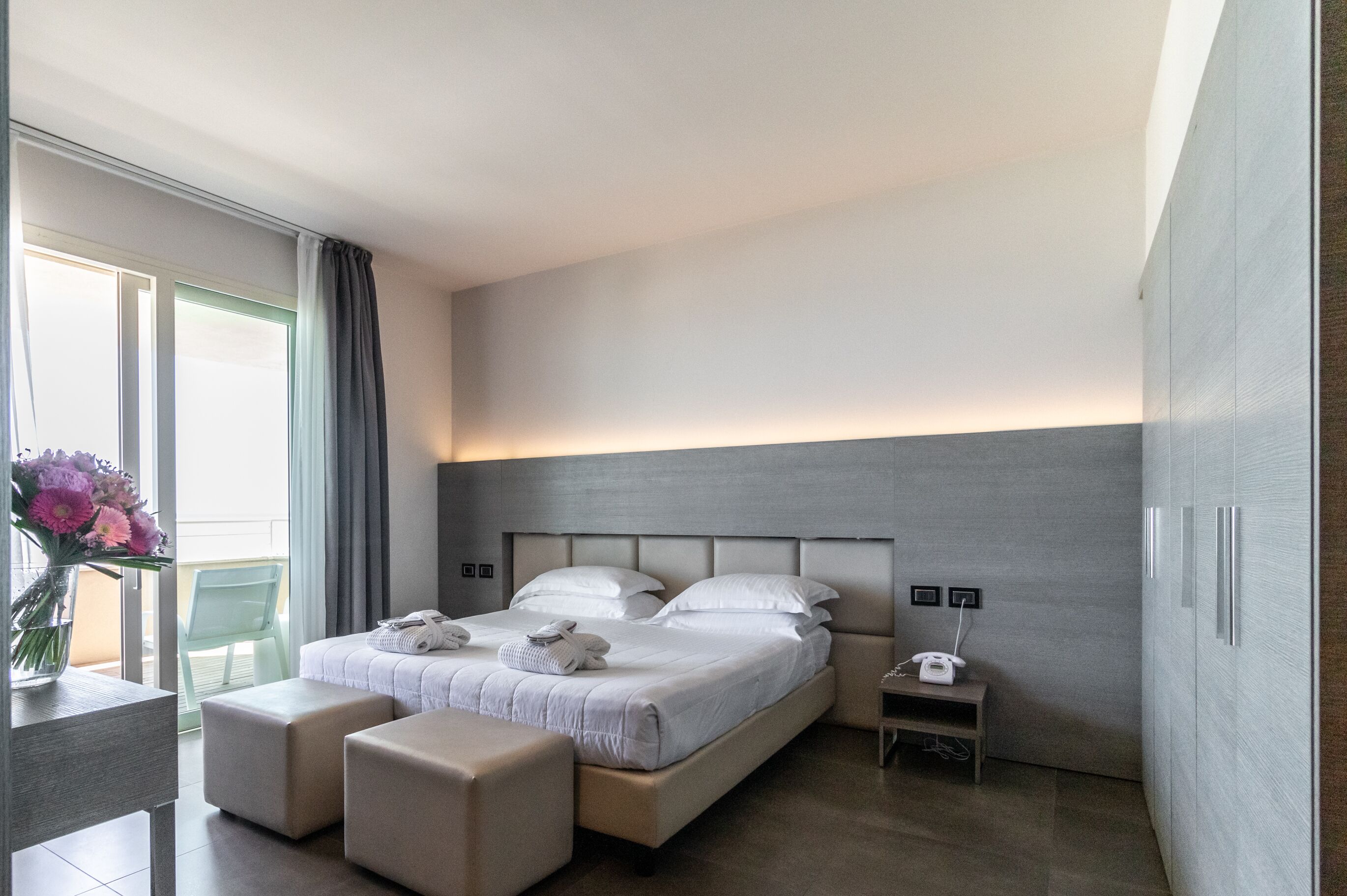 A modern hotel room with a large bed, two bedside tables, a small desk, and a chair. There is a window with curtains, a vase of flowers, and a view of the outside.