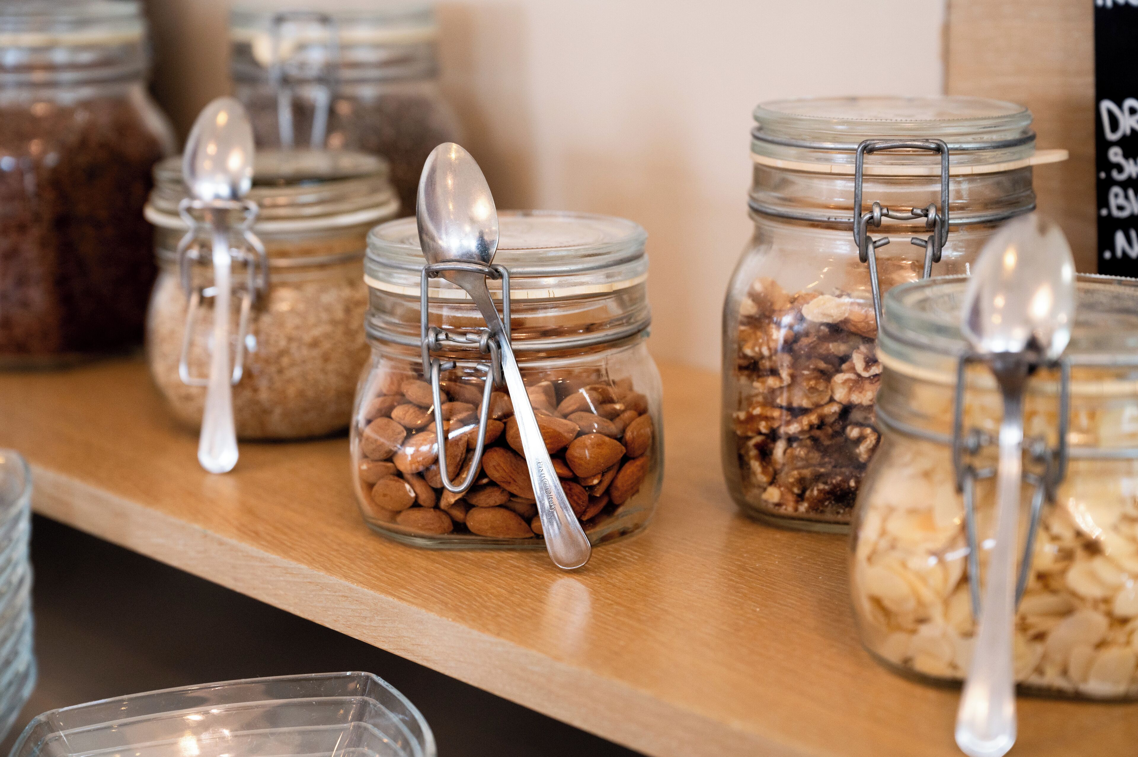 A shelf with glass jars containing various nuts and grains, each with a spoon.