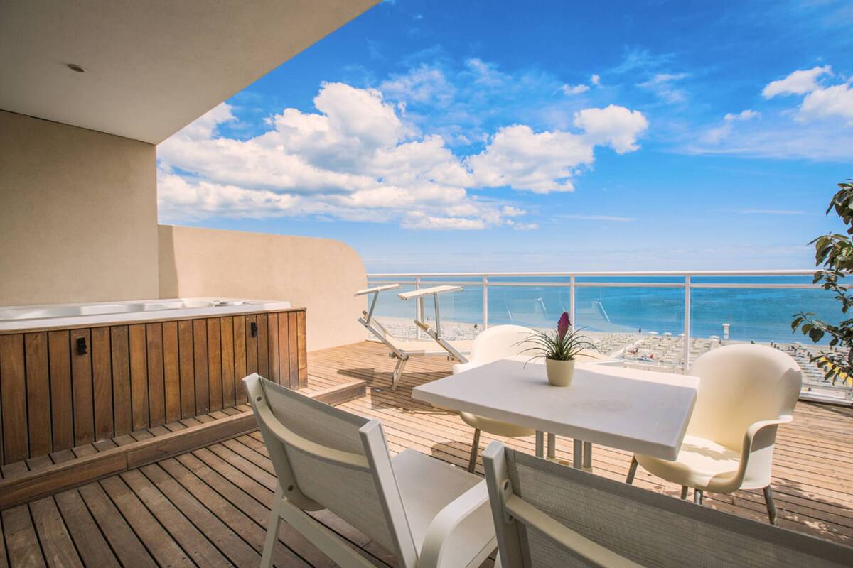 Eine Holzterrasse mit Tisch, Stühlen, einem Whirlpool und Blick auf Strand und Meer.