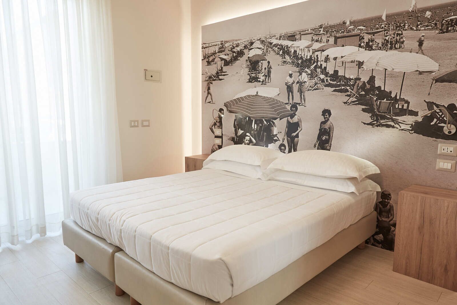 A modern hotel room with a large bed, a mural of a beach scene, and a large window with sheer curtains.