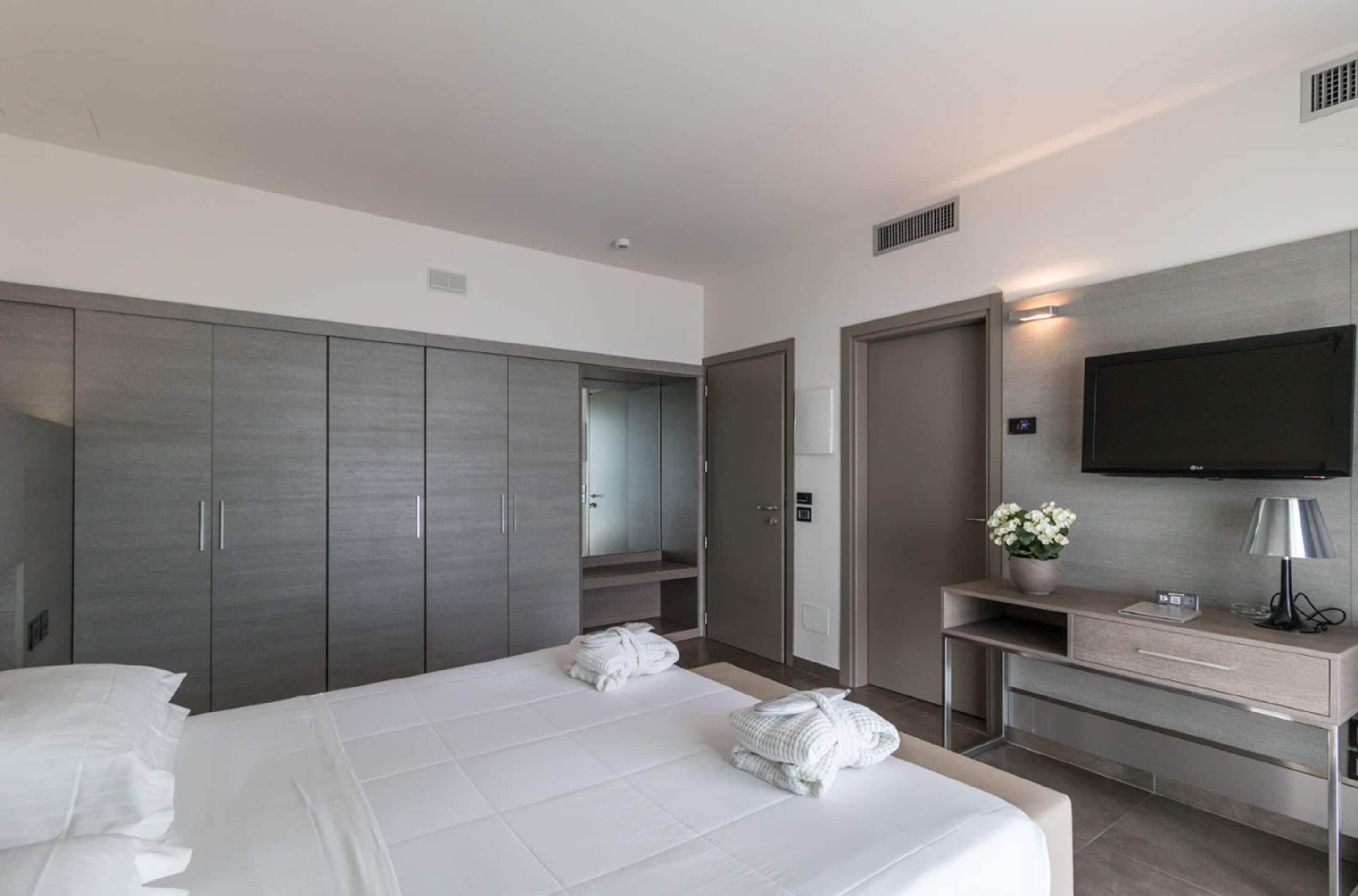 A modern hotel room with a large bed, grey wardrobe, a TV mounted on the wall, and a small desk with a lamp and flowers.