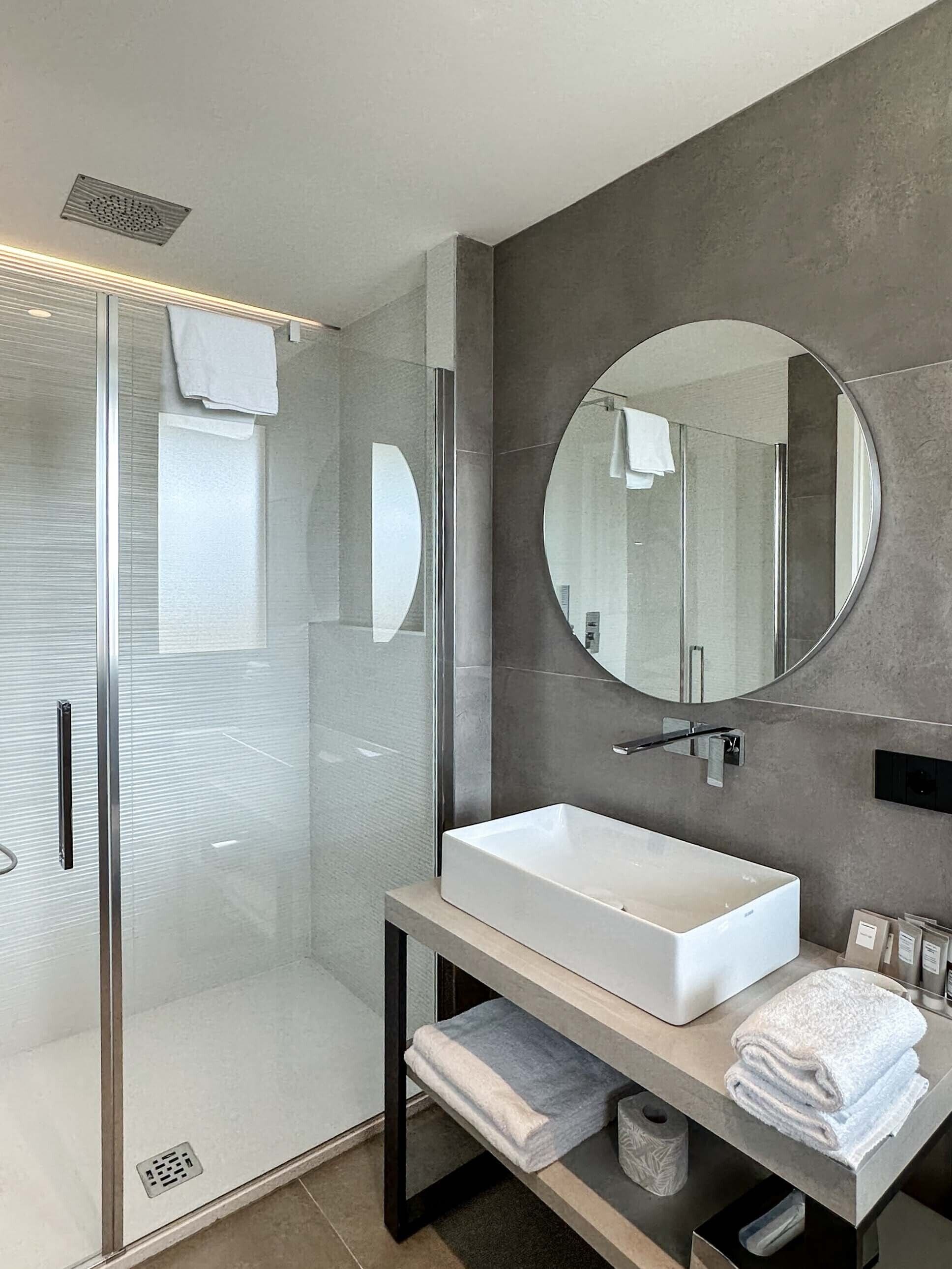 A modern bathroom with a glass-enclosed shower, a large round mirror, and a white sink.