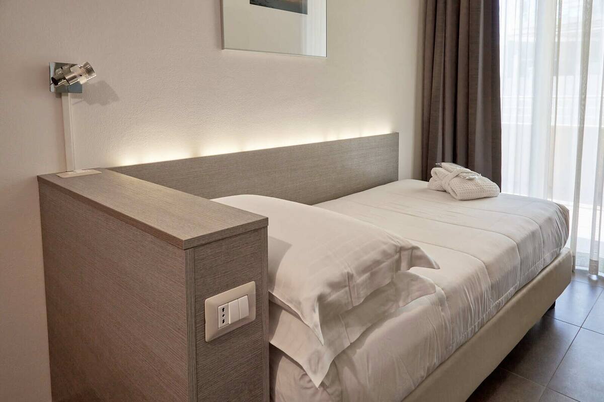 Ein ordentlich bezogenes Bett mit weißen Leinen, ein Nachttisch mit Steckdose und ein an der Wand befestigter Spiegel.