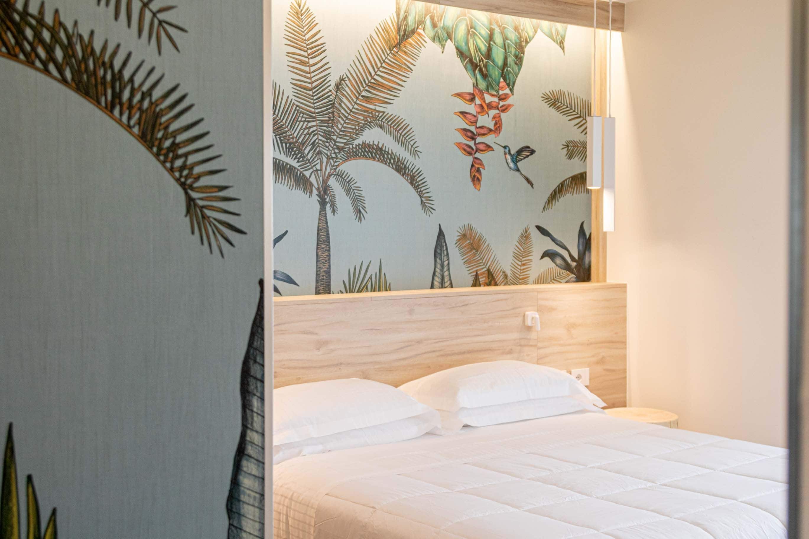 Quarto com cama, roupa de cama branca, cabeceira de madeira e parede com padrão de folha tropical.