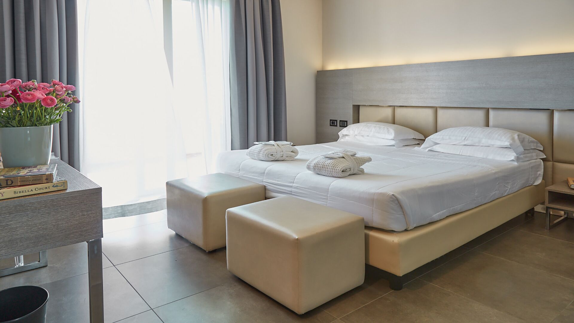 A modern hotel room with a large bed, two bedside tables, a bedside lamp, and a view of the outdoors.