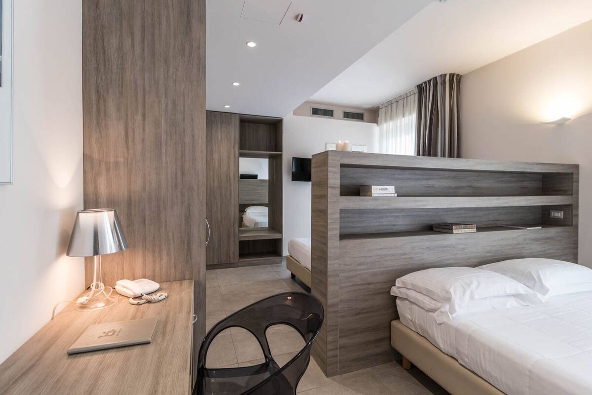 Ein modernes Hotelzimmer mit einem Bett, einem Schreibtisch mit einem Laptop, einem Stuhl, einem Kleiderschrank und einem fest eingebauten Regal mit Büchern.