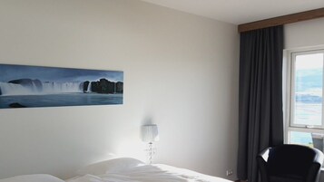 A hotel room with a bed, a lamp, a chair, a window with curtains, and a painting of a waterfall.