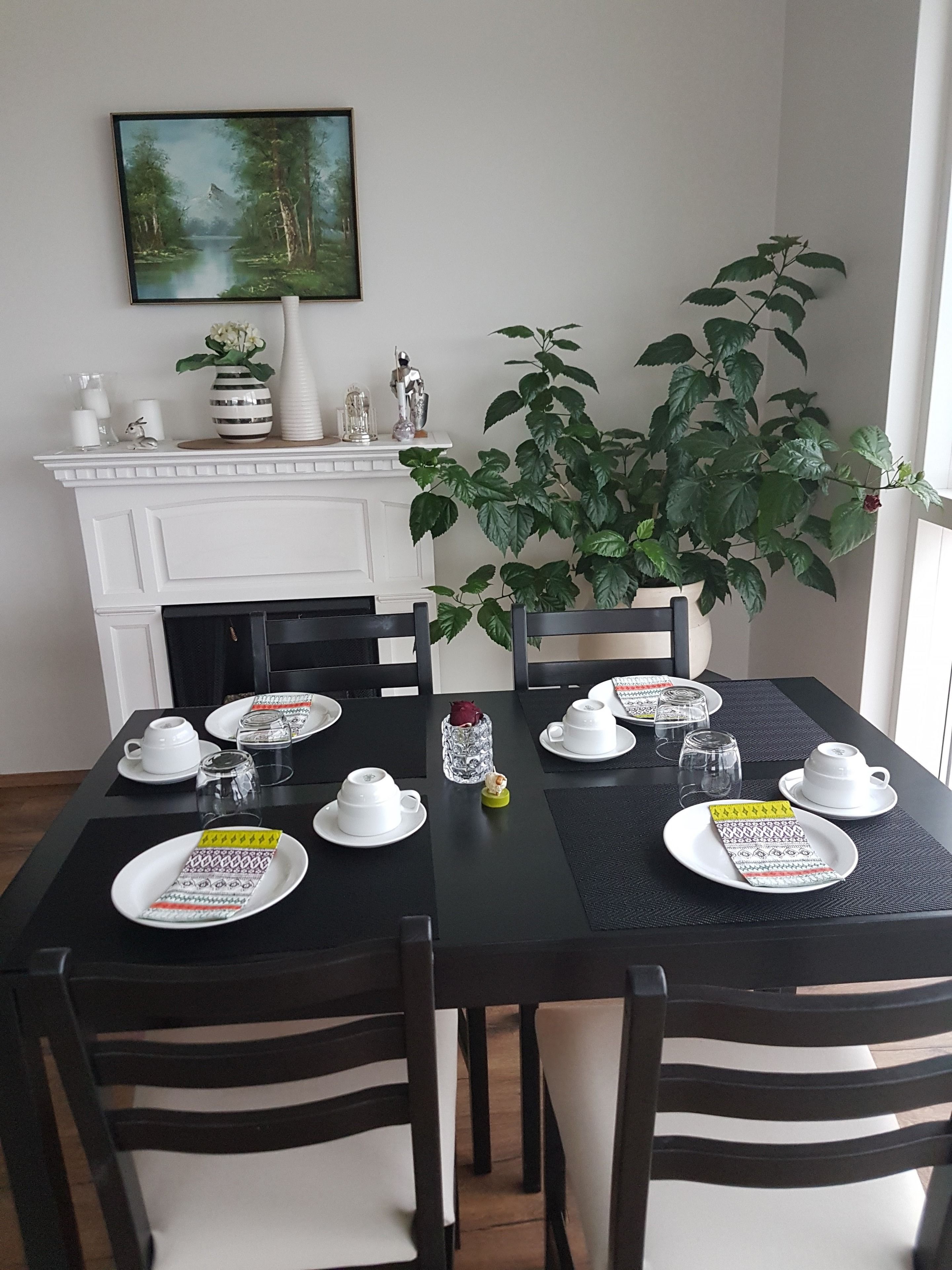 Mesa de comedor lista para cuatro personas, con platos, tazas y vasos blancos, una planta en maceta y una chimenea al fondo.