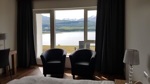 A hotel room with a large window offering a view of a lake and mountains. There are two black chairs and a desk with a lamp. - Hafdals Hotel (Akureyri)