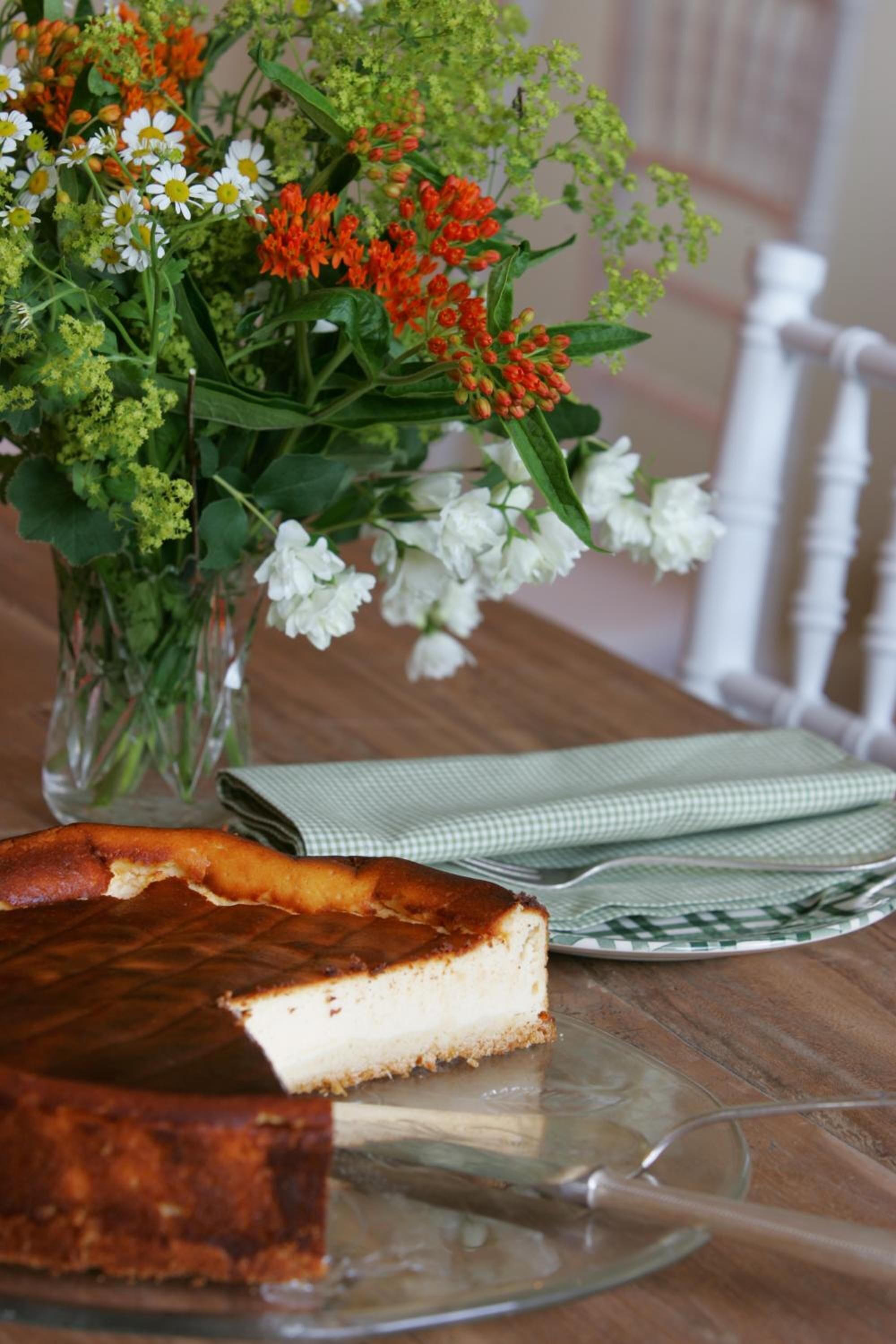 A cheesecake on a glass plate with a checkered napkin beside it, a vase of flowers, and a white chair in the background.