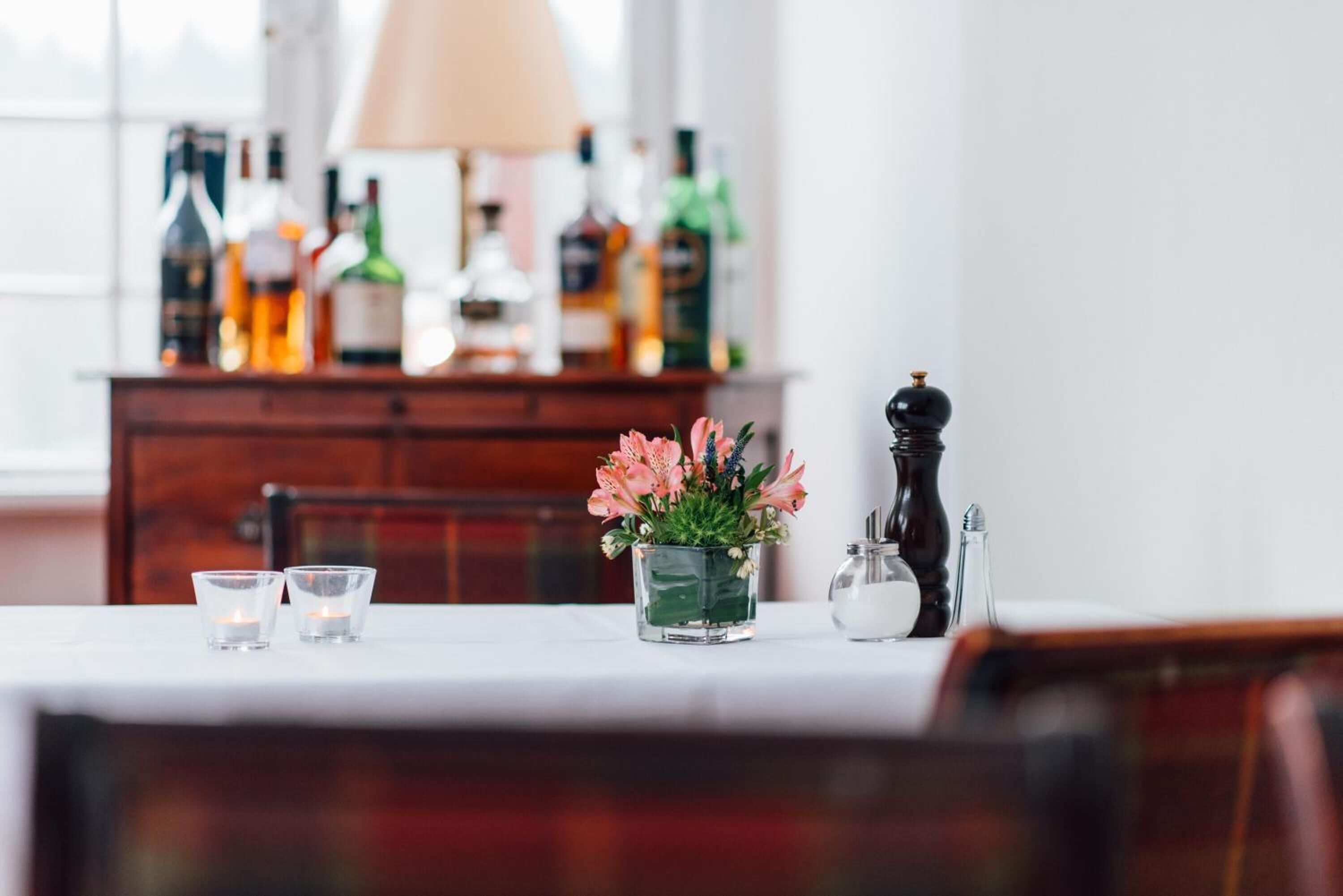 Un tavolo da pranzo con un vaso di fiori, saliera e pepiera, e due bicchieri. Sullo sfondo, un mobiletto in legno con bottiglie e una lampada.