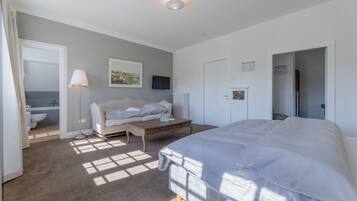 Un dormitorio con una cama grande, un sofá, una mesa de centro y un televisor. Se ve un baño al fondo.
