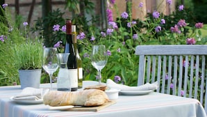 A table set outdoors with a bottle of wine, glasses, a loaf of bread, and a small bowl, surrounded by greenery and flowers.