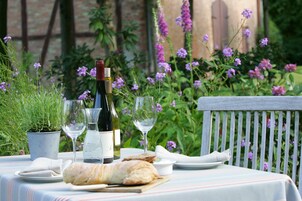 A table set outdoors with a bottle of wine, glasses, a loaf of bread, and a small bowl, surrounded by greenery and flowers.