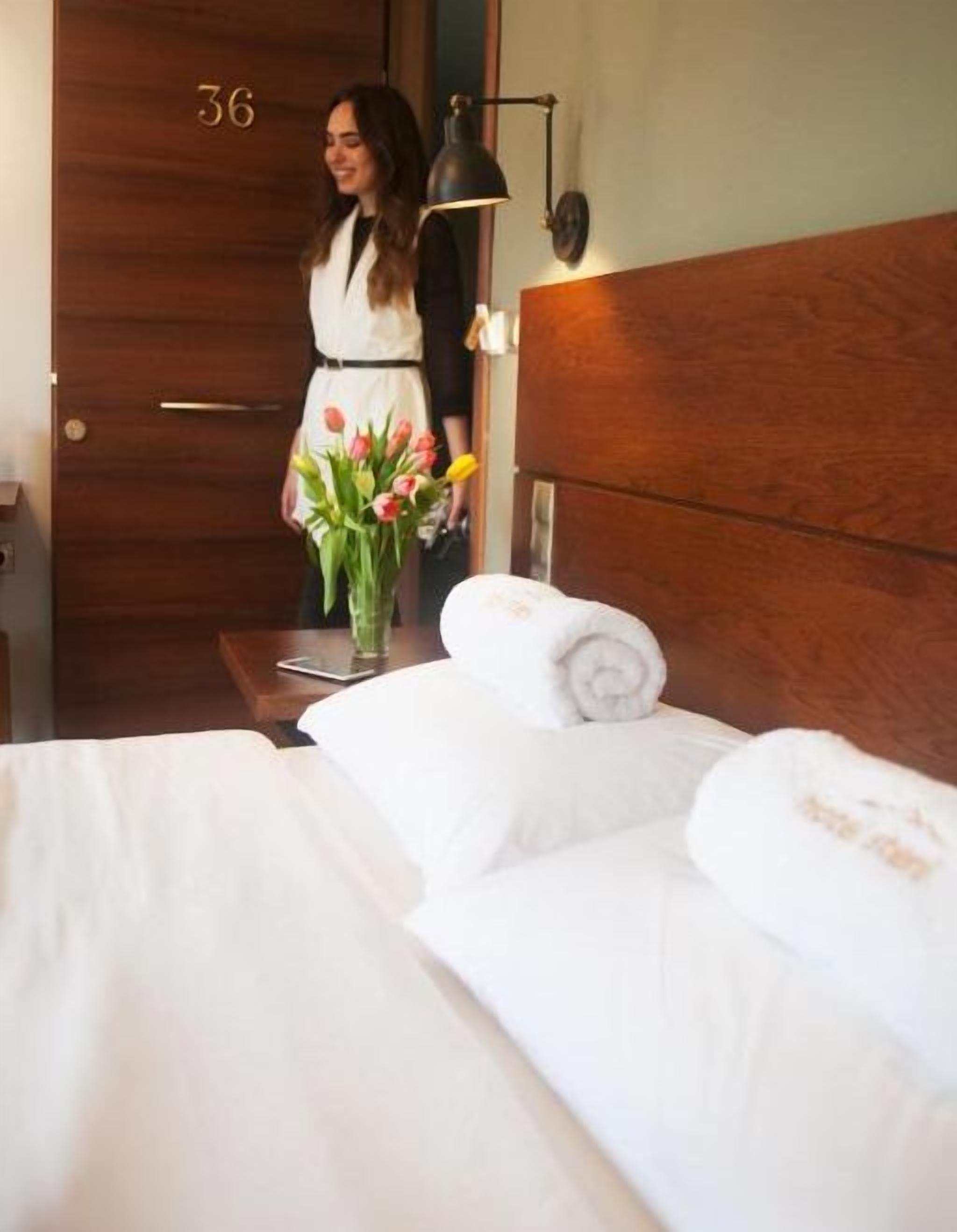 A hotel room with a wooden headboard, a bed with white linens, a nightstand with a lamp, and a vase of tulips.