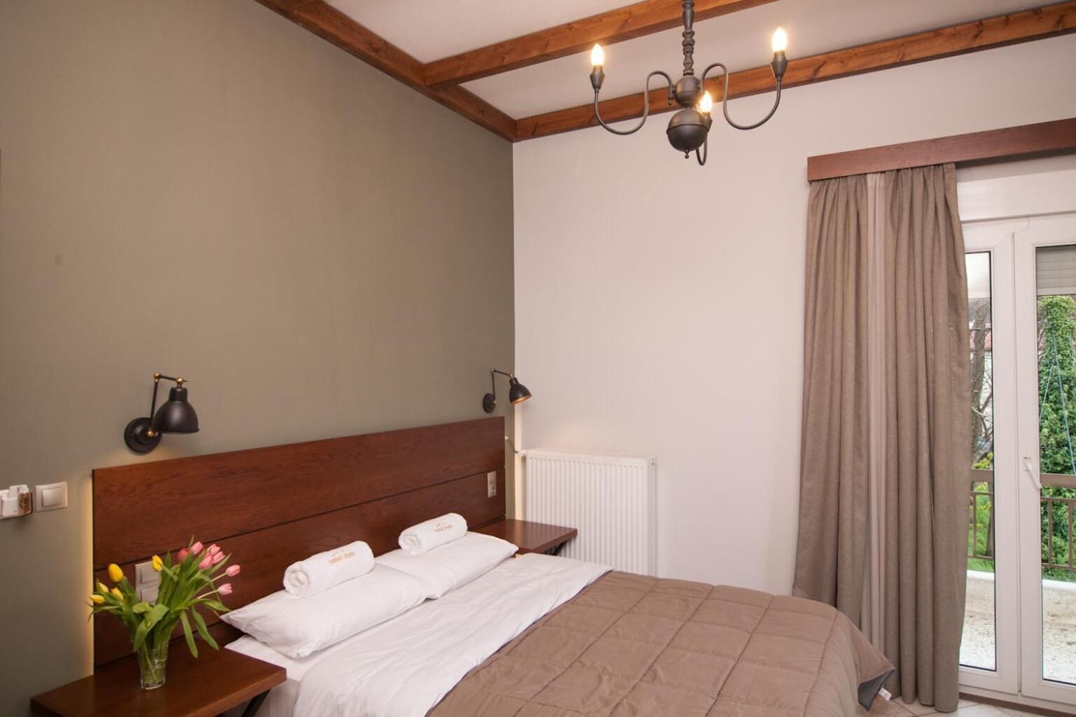 A bedroom with a wooden headboard, a large bed with white linens, a bedside table with a lamp, and a view of greenery through the window.