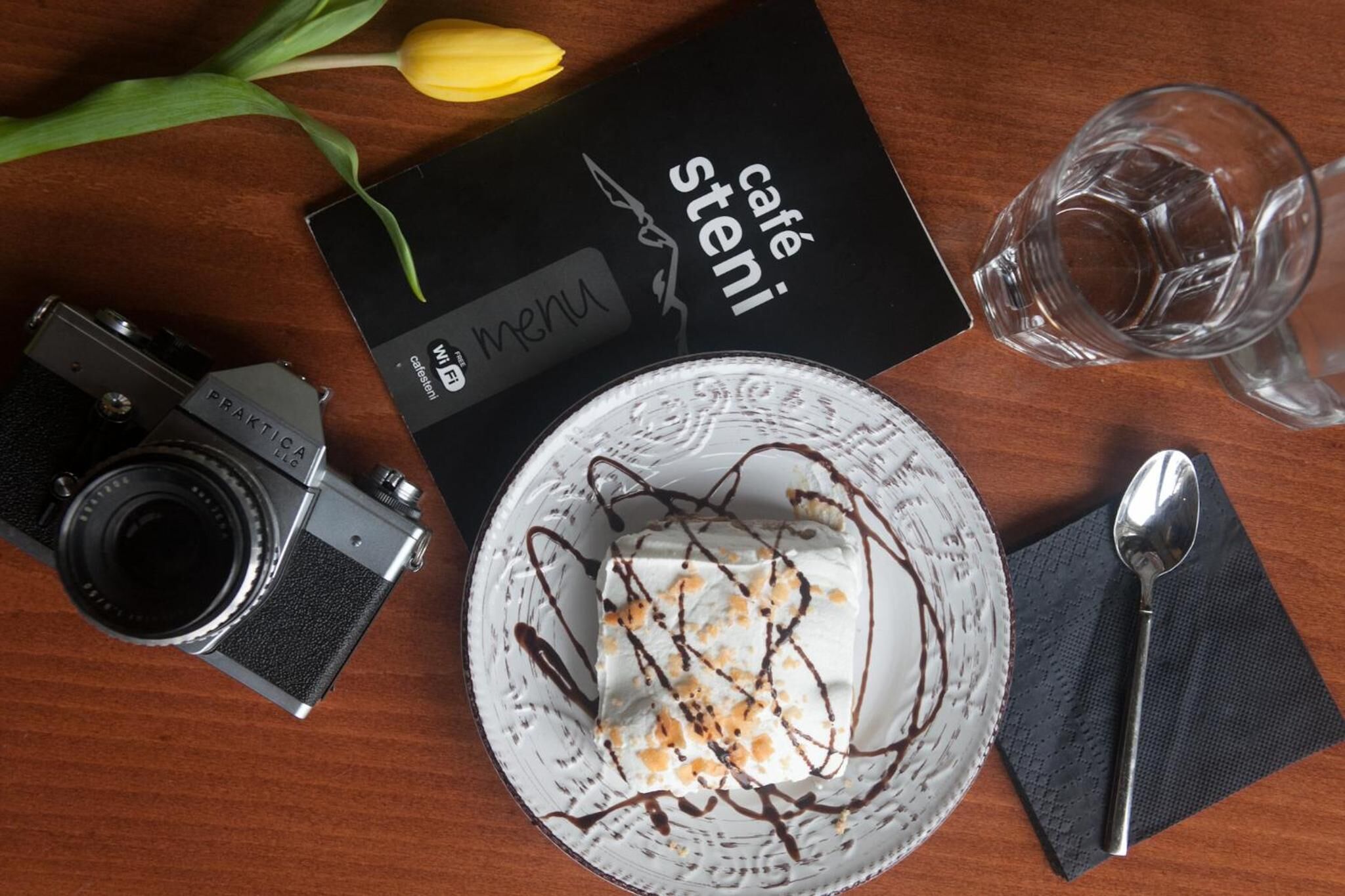 Ein Desserteller mit Schlagsahne und Schokoladenglasur, eine Vintage-Kamera, eine Speisekarte und ein Glas Wasser auf einem Holztisch.