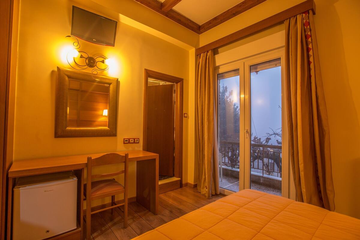 A hotel room with a bed, a desk with a chair, a small refrigerator, a television mounted on the wall, and a sliding glass door leading to an outdoor area.