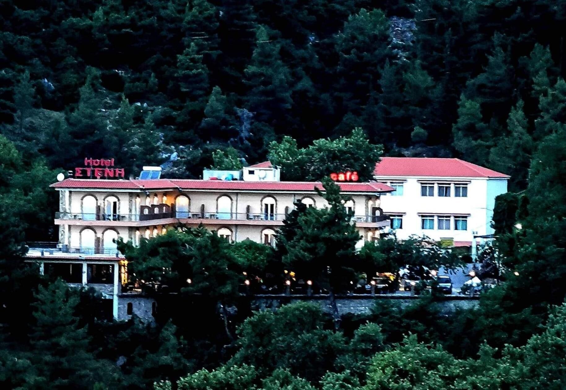 Un edificio de dos pisos con techo rojo, un letrero que dice "Hotel ETENH" y un letrero de cafetería. Está rodeado de un bosque frondoso.