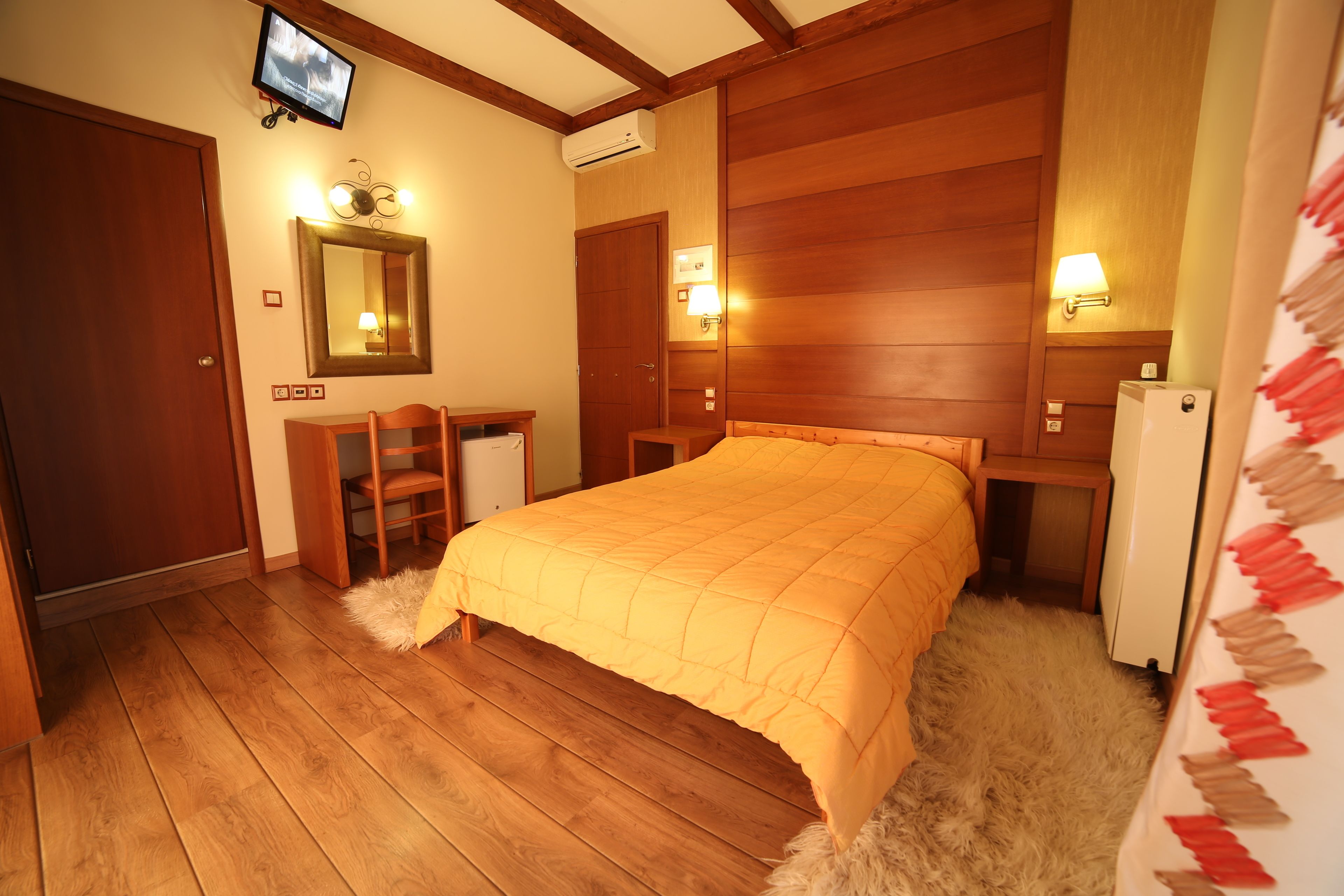 Quarto com cama de madeira, escrivaninha, cadeira, espelho e televisão fixada na parede.