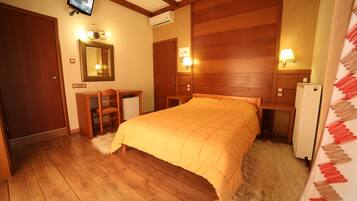 Quarto com cama de madeira, escrivaninha, cadeira, espelho e televisão fixada na parede.