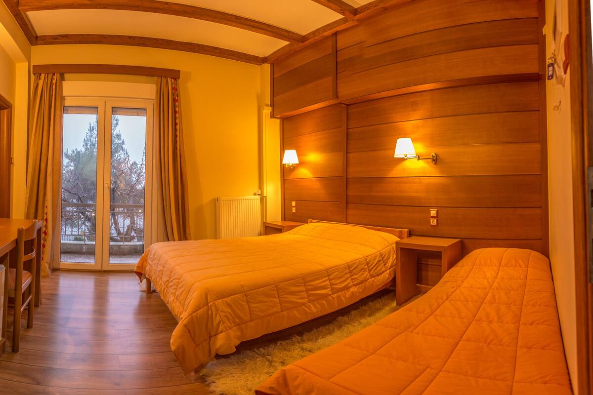 A room with two beds, wooden headboard, two bedside lamps, a wooden desk, and a view through a glass door.