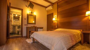 Un dormitorio con cama de madera, mesita de noche, escritorio y espejo. Se ve una puerta que conduce a un baño con inodoro y cortina de ducha.