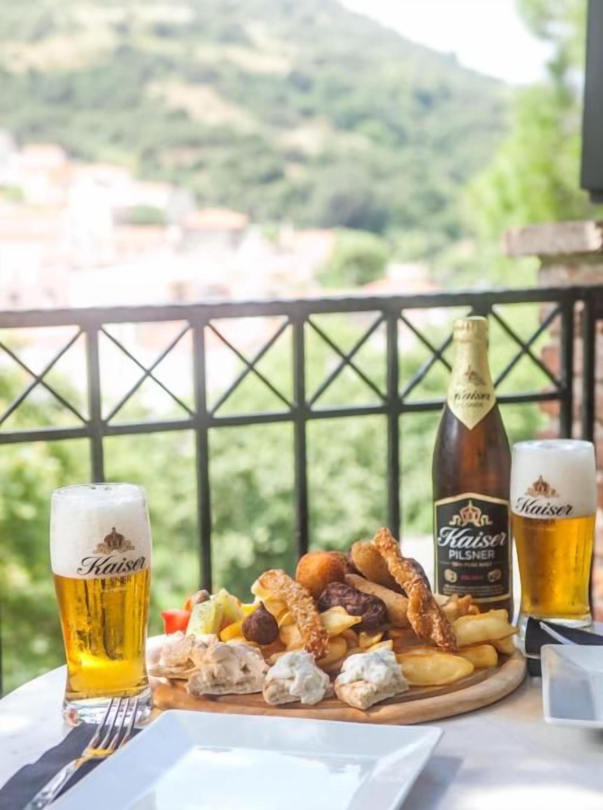 A table with a plate of food, two glasses of beer, and a bottle of beer on a balcony with a scenic view.
