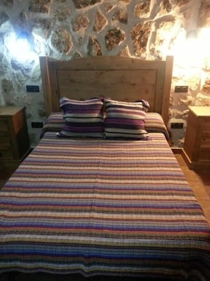 Ein Bett mit gestreifter Decke und zwei Kissen, ein Holz-Kopfschild und eine Steinsäule im Hintergrund.